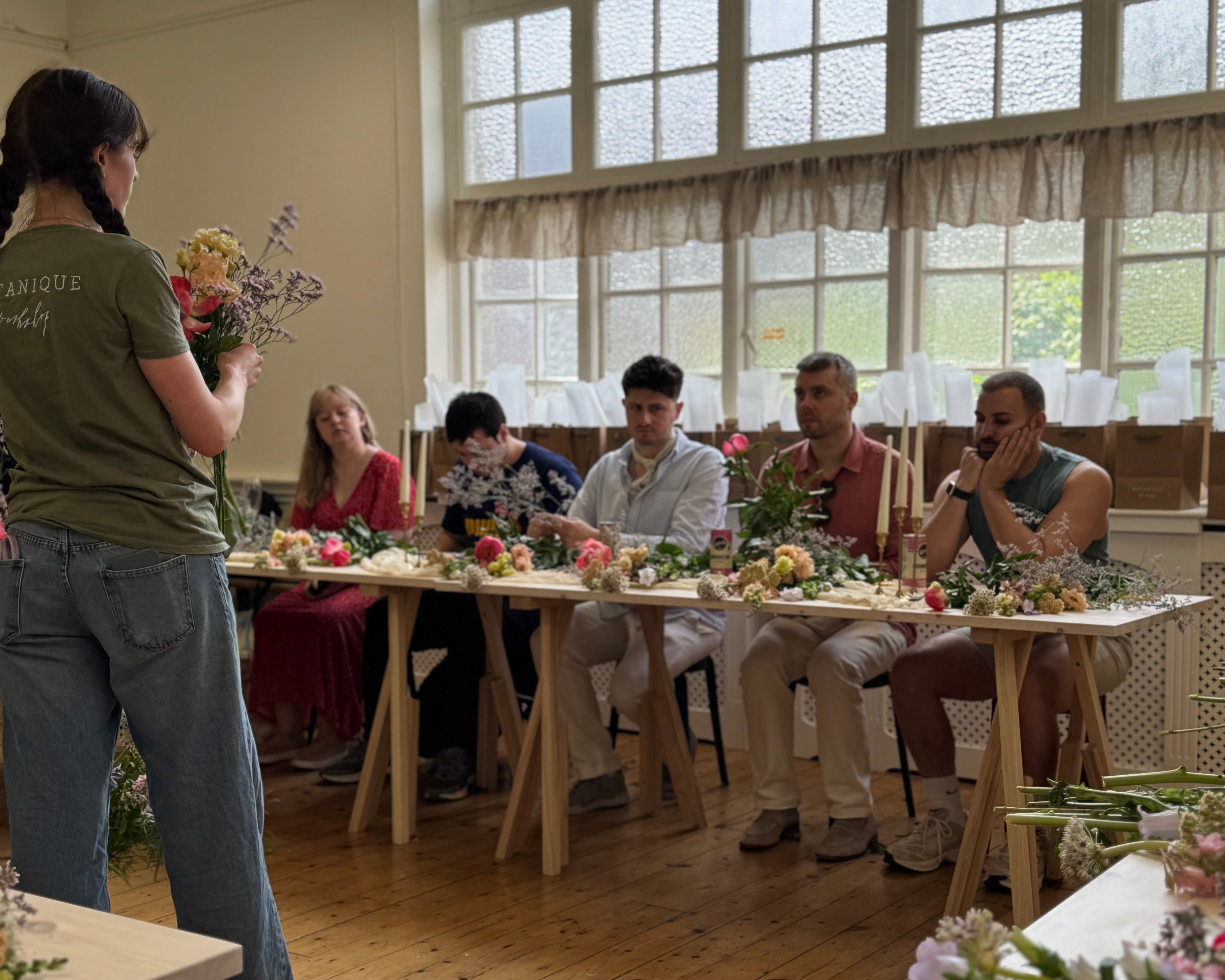 Group of people sitting at tables following a florist at a flower workshop in london, in a room with large windows.