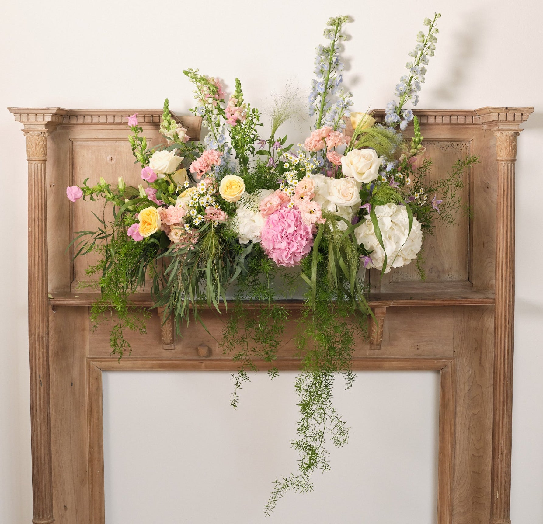 A large wedding trough flower arrangement filled with an overflow of pastel colored flowers, including shades of pink, blue, yellow, and white, displayed on a wooden mantlepiece.
